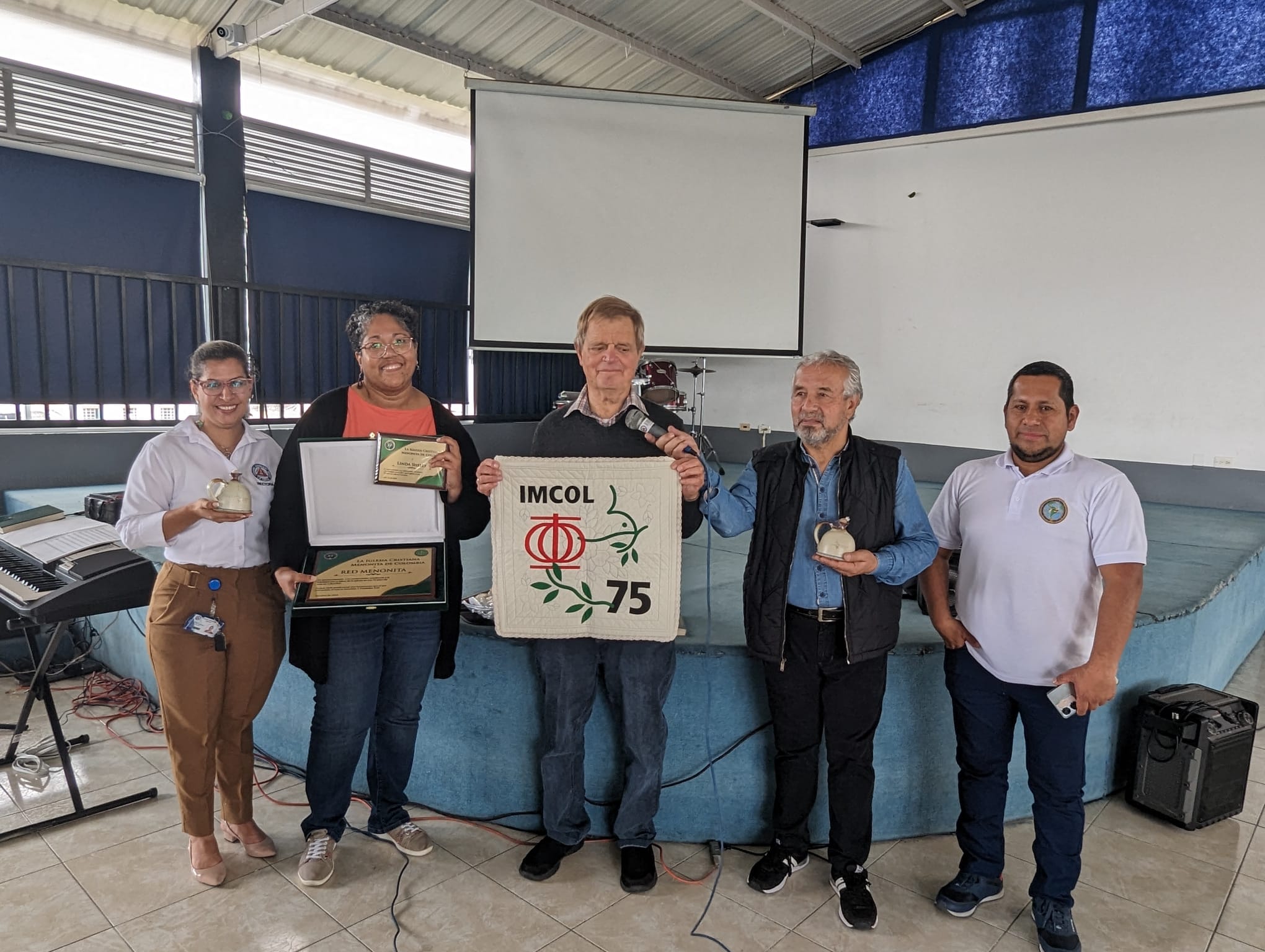 IMCOL administrator Oscar Ospina, President Carlos Moreno and pastor Peter Stucky (right to left) receive a quilted wall hanging as a 75th anniversary gift from Mennonite Mission Network. It is an artistic integration of the IMCOL logo (taken from the former General Conference Mennonite Church) and the Mission Network dove. Colegio Americano Menno Director Rebeca Muñoz and Mission Network Interim Executive Director Marisa Smucker (left to right) display other gifts. Photo by Martin Gunawan.
